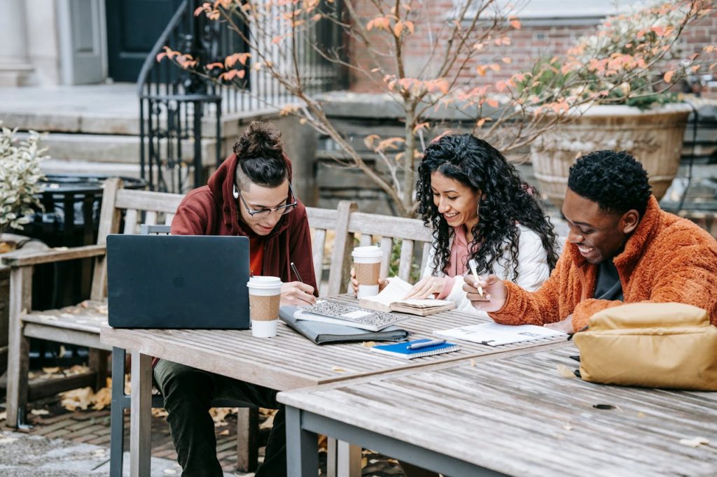 Group of positive multiracial classmates sitting at wooden table with takeaway coffee and laptop while writing in copybooks during studies on street near building