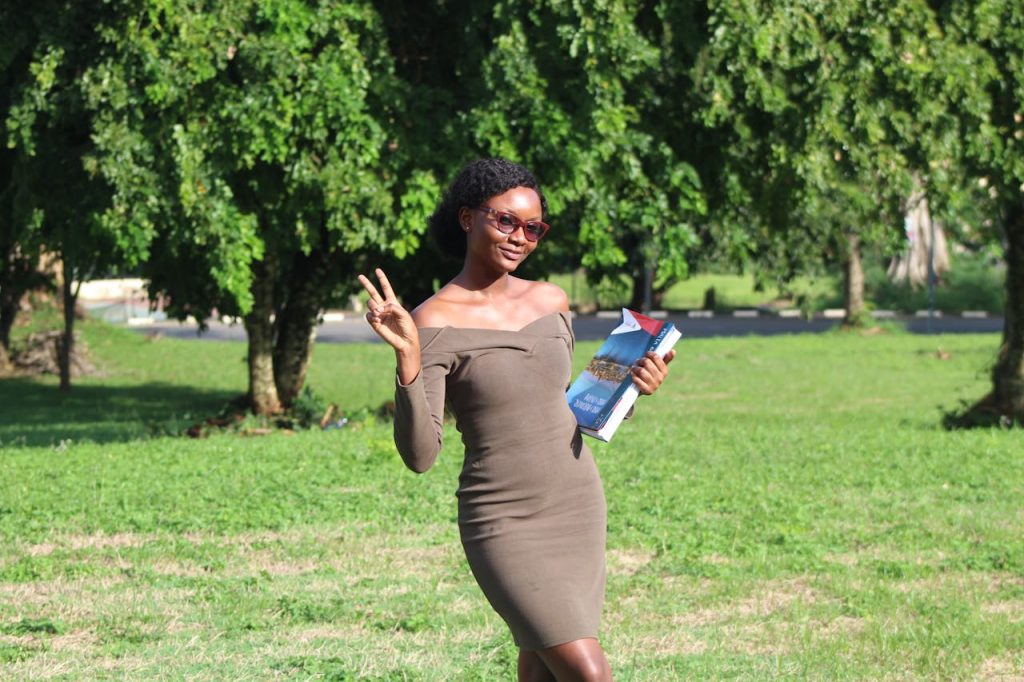 Stylish woman holding books and peace signing in a sunny Kumasi park.