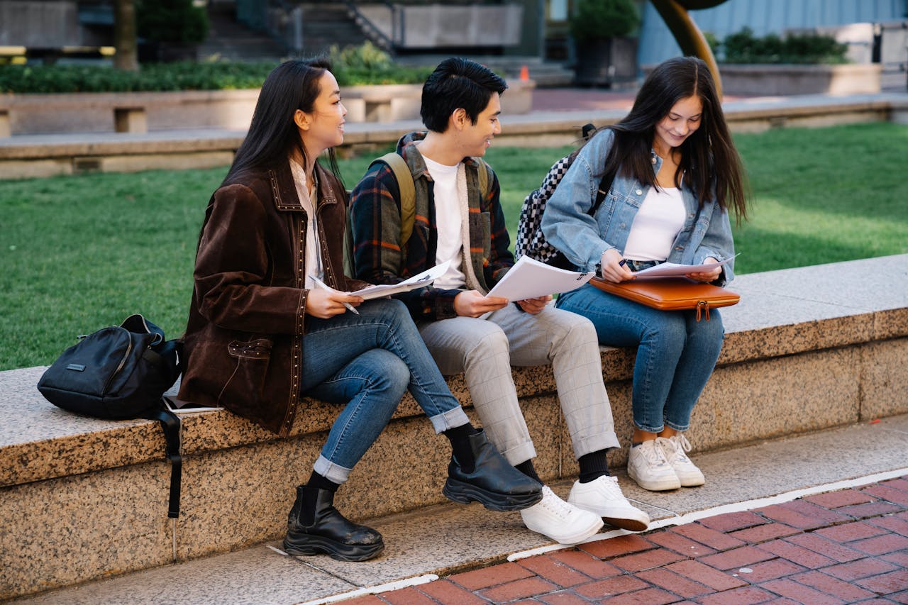 Three college students engaged in study session on a campus bench, enjoying a sunny day.